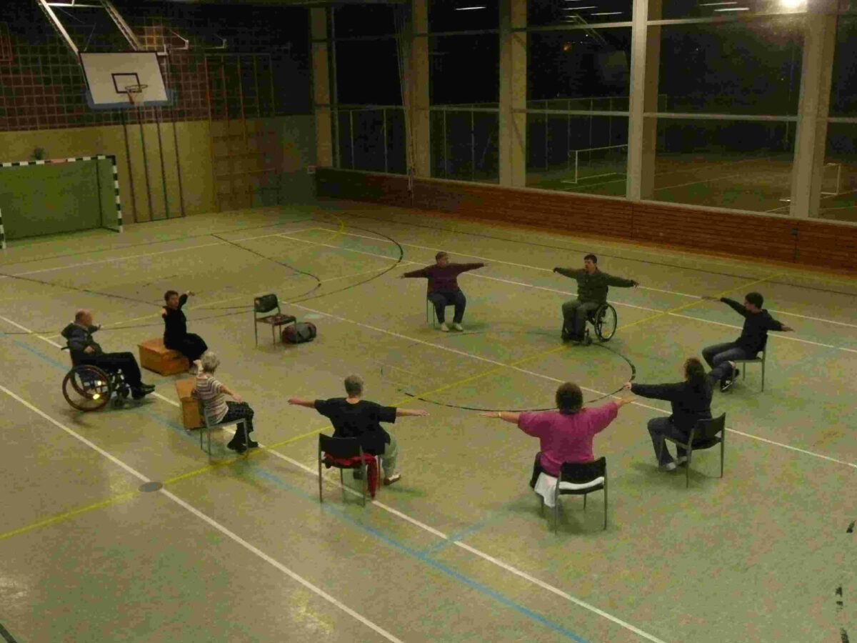 Das Bild zeigt eine Gruppe Rollstuhlfahrer und Fußgänger auf einem Stuhl sitzend bei der Gymnastik.